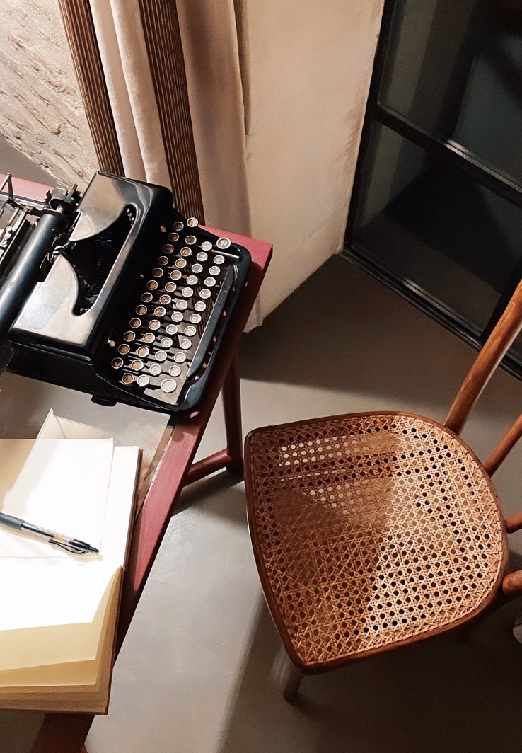 A typewriter on a small desk beside a cane chair