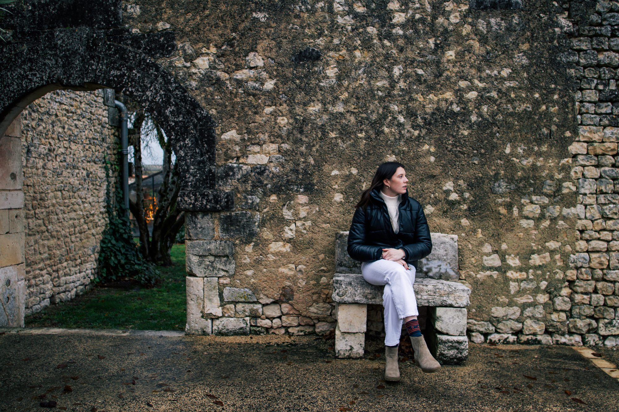 A person seated on a stone bench before an old stone wall with an arched opening