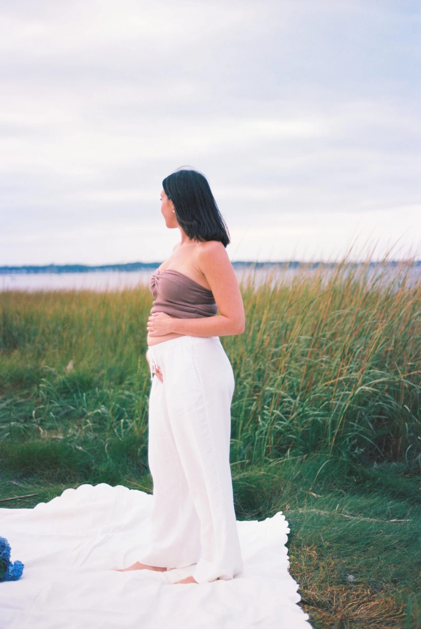 A woman in a long white skirt standing in tall grass by the water